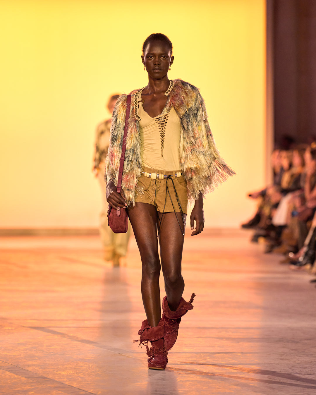 Feminine model on the catwalk, dressed in the Noela short jacket with feather-effect bangs, the Sakina top in draped faux suede, camel shorts, the Rosly jewel belt in pearls and the Winki boots in suede and braided leather in burgundy.