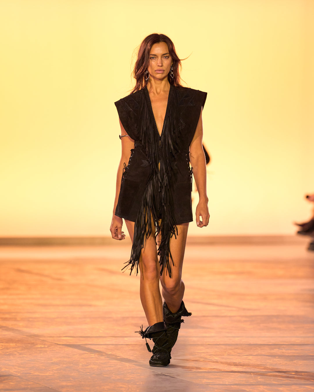 Female model on the catwalk, dressed in a black fringed suede sleeveless vest, black shorts and black suede and braided Winki boots.
