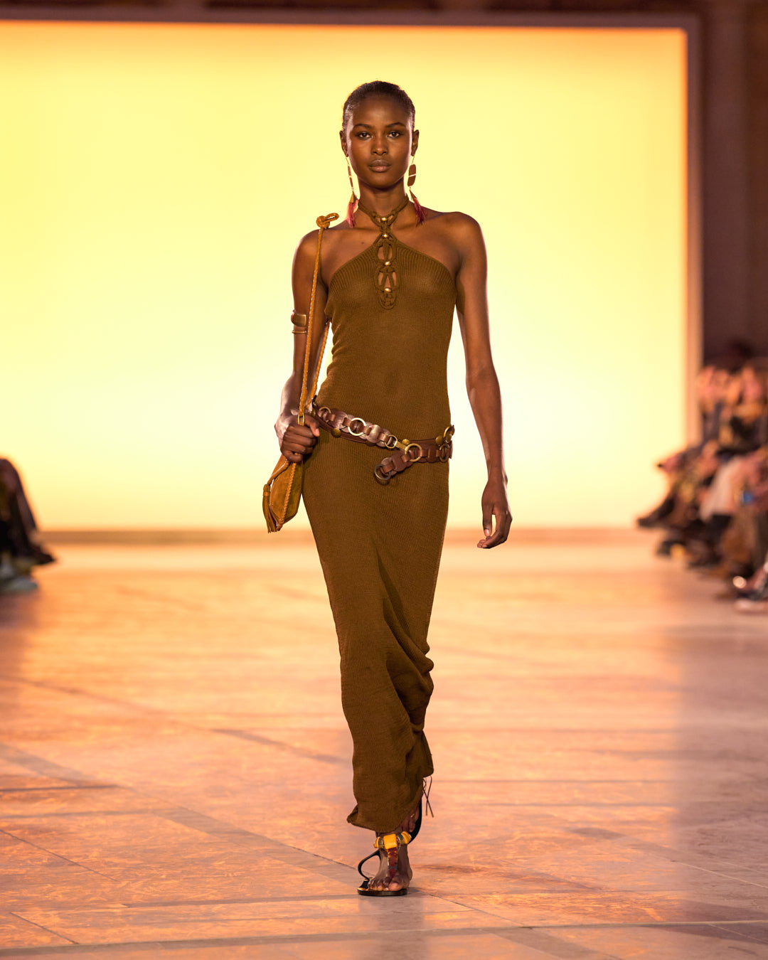 Female model on the catwalk, wearing the Eileen Long Dress in fine bronze mesh, and the cognac-colored Anahi bag.