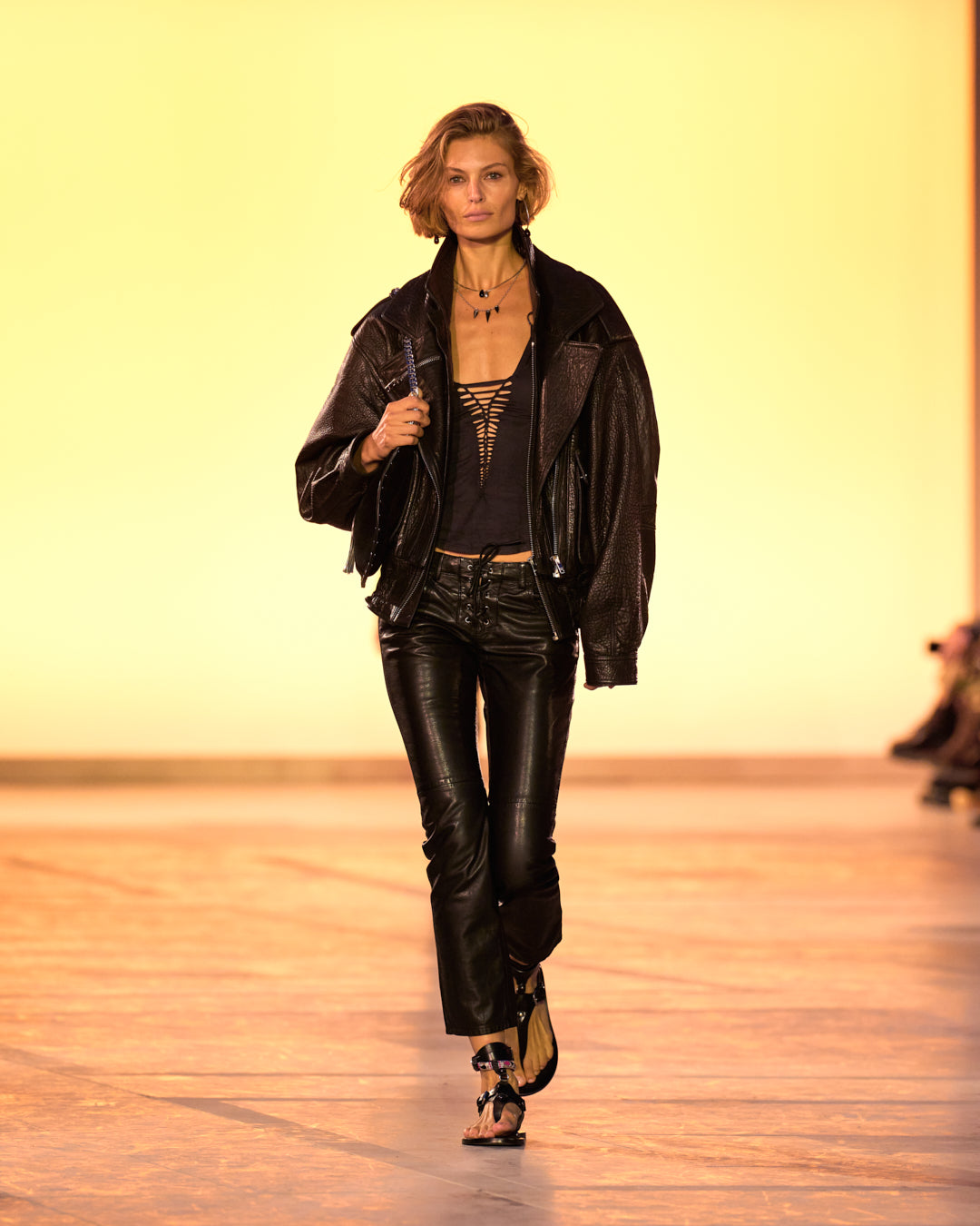 Female model on the catwalk, wearing the Aneli pocket jacket in lambskin, Sakina top in faux suede draped in black, Vanel lace-up denim jeans in black and black sandals.