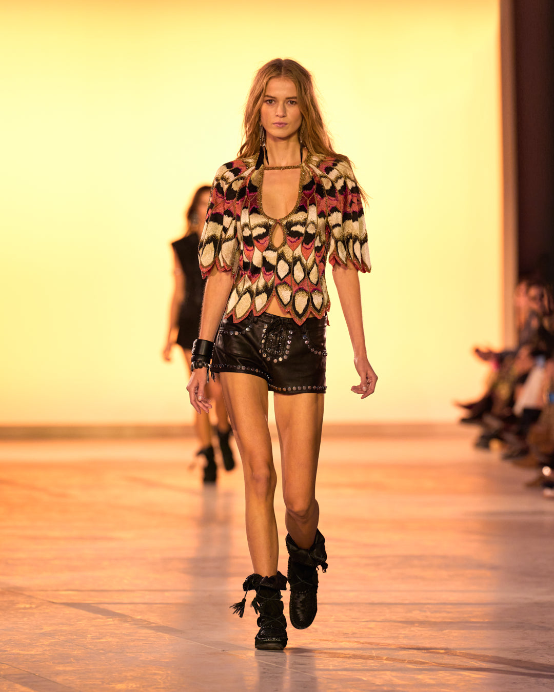 Female model on the catwalk, wearing the Aynia top in butterfly embroidered silk, the black Elsanna lace-up shorts in studded leather and the Winki boots in black suede and braided leather.