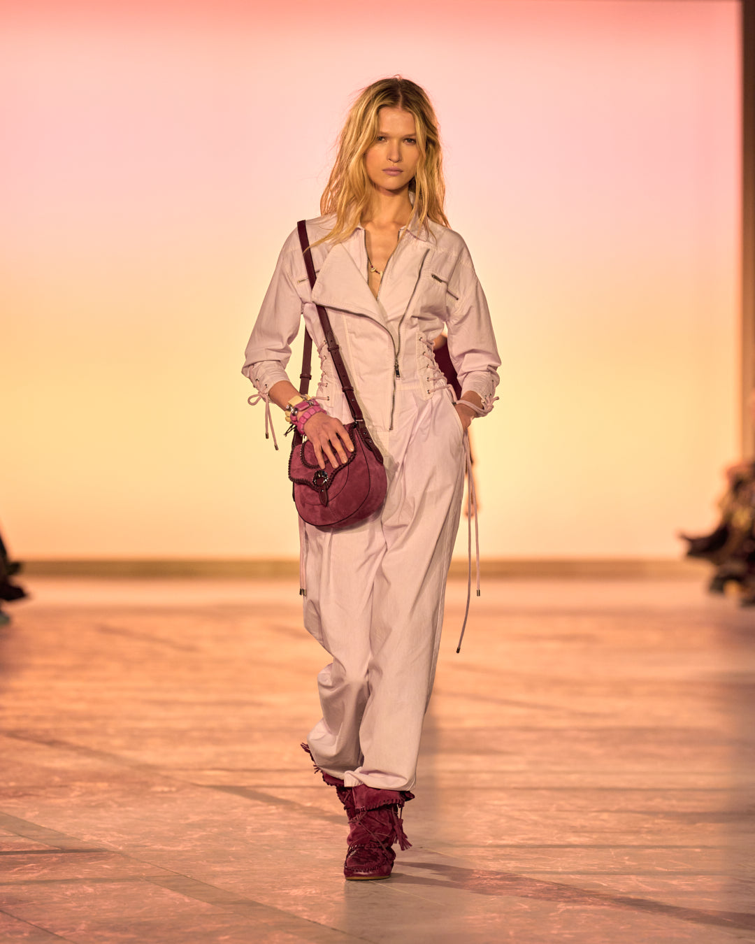 Female model on the catwalk, dressed in a pink jumpsuit, a raspberry altay small bag and burgundy suede and braided Winki boots.